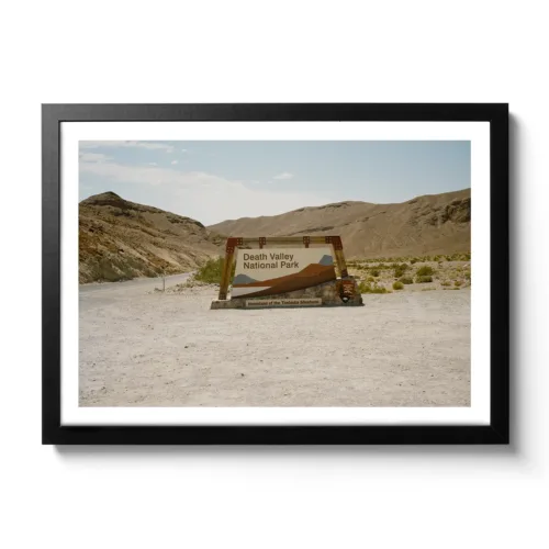 Bildaufnahme eines Schotterparkplatzes, mit dem Einfahrtsschild zum Death Valley National Park