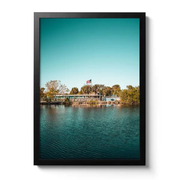 Landschaftsaufnahme von Captain Jack’s Airboat Tour in den Everglades am Wasser, umgeben von Bäumen, mit einer amerikanischen Flagge im Hintergrund unter klarem, blauem Himmel.