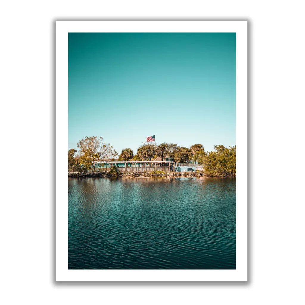 Landschaftsaufnahme von Captain Jack’s Airboat Tour in den Everglades am Wasser, umgeben von Bäumen, mit einer amerikanischen Flagge im Hintergrund unter klarem, blauem Himmel.