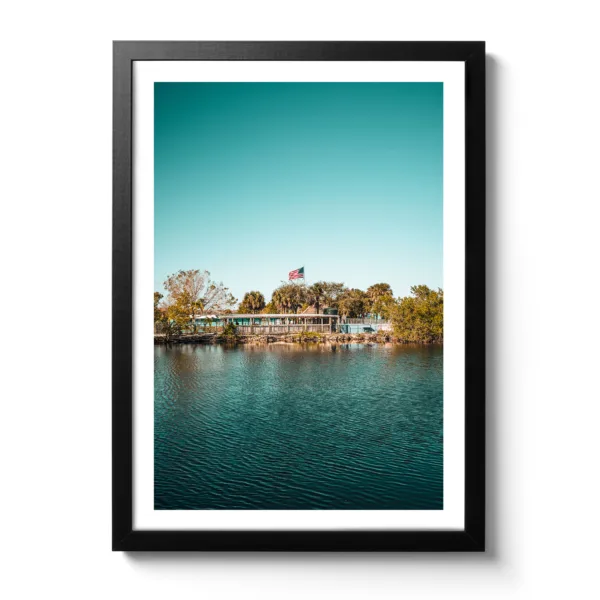 Landschaftsaufnahme von Captain Jack’s Airboat Tour in den Everglades am Wasser, umgeben von Bäumen, mit einer amerikanischen Flagge im Hintergrund unter klarem, blauem Himmel.