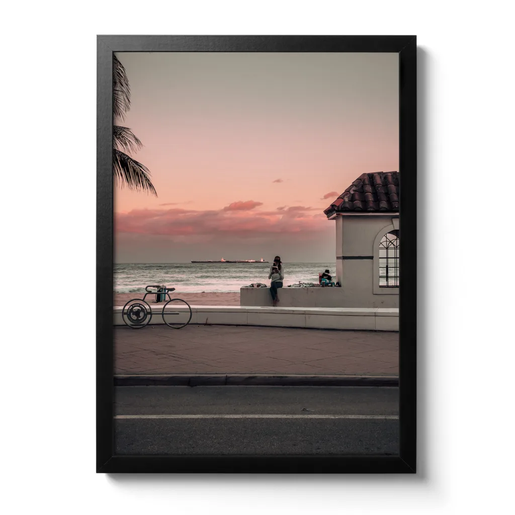 Bildaufnahme eines Sonnenuntergangs, wo eine Frau am Randstein sitzt und ein Buch liest. Im Hintergrund befindet sich das Meer und eine kleine weiße Hütte.